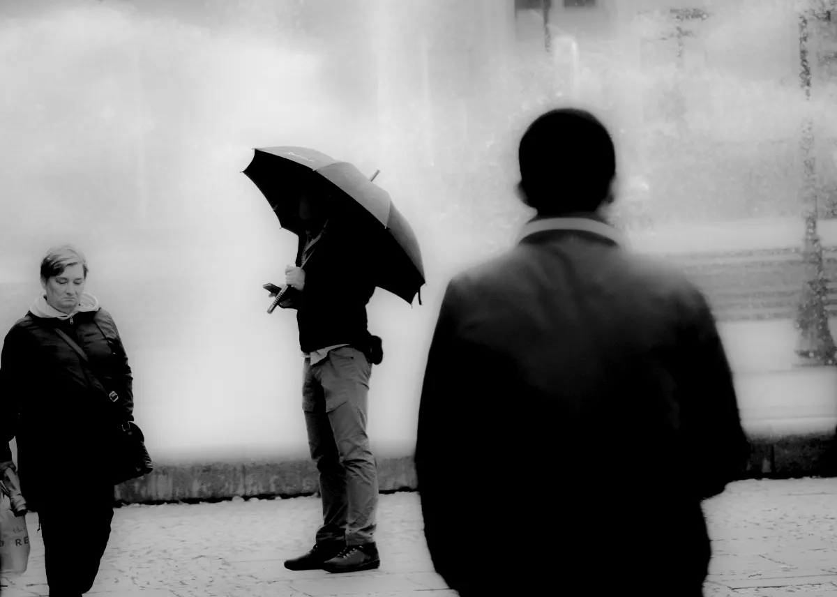 Man checking his phone beneath a black umbrella by a fountain